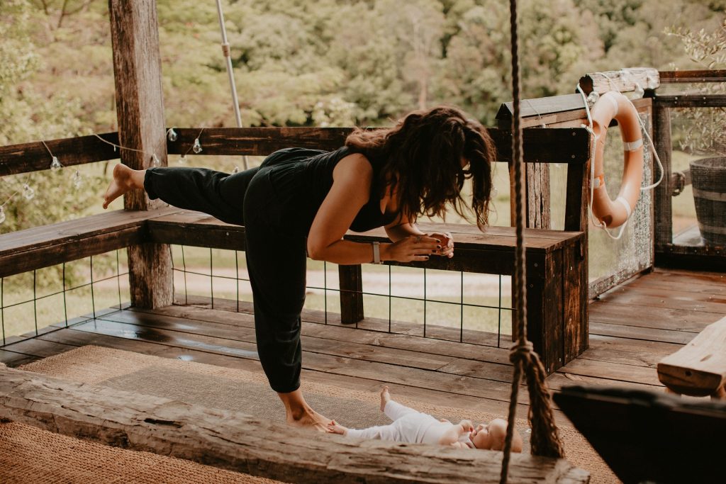 A woman performs Warrior 3 while looking at a baby